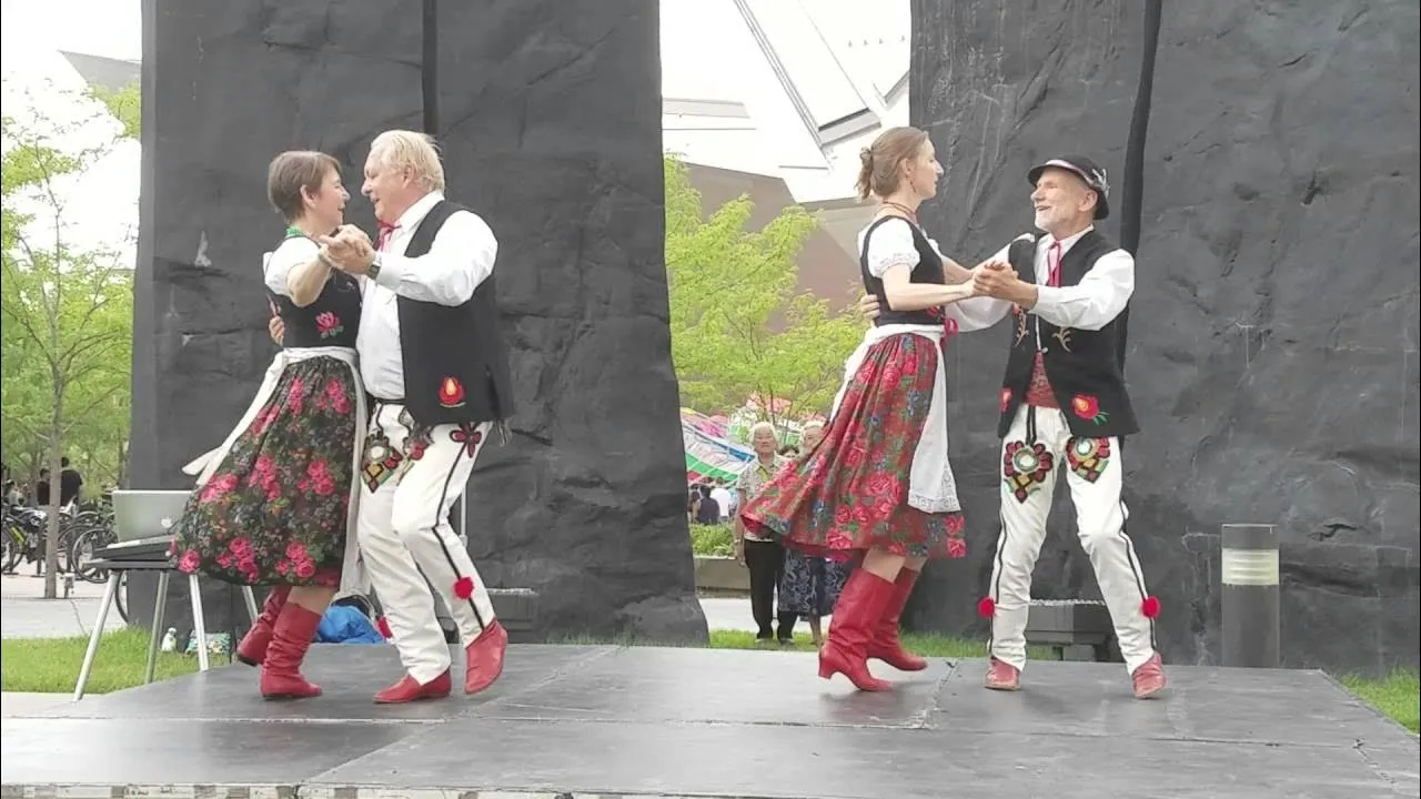 Two pairs of postoley folk dancers dancing on a stage in a park.